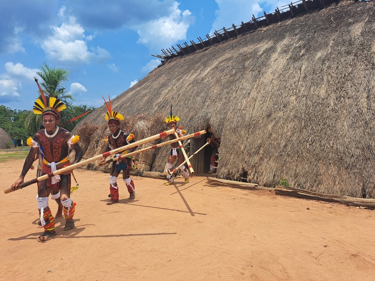 #PraCegoVer: Foto em ambiente externo mostra indígenas Kuikuro saindo de uma grande construção tradicional de palha na Aldeia Afukuri, no Xingu. Com grafismos corporais, cocares coloridos e adornos nos braços e pernas, carregam longas lanças de madeira enquanto caminham. Ao fundo, a estrutura coberta de palha está cercada por vegetação, sob um céu azul com nuvens (Divulgação/Vivalá) - expedições BioConexão